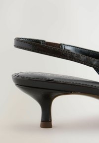 Svart högklackad sandal med en smal rem, som visar en slät sula och en lätt böjd klack, placerad mot en ljus bakgrund.