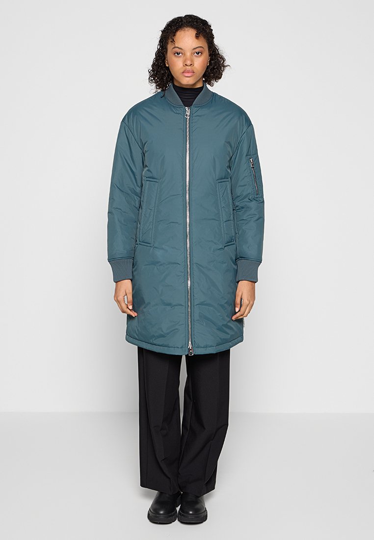 Türkisfarbene Daunenjacke mit Reißverschluss vorne, gerippten Bündchen und Seitentaschen. Das Model trägt sie über dunklen, weiten Hosen und schwarzen Schuhen.