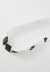 Cinghie in tessuto bianco con loghi Nike neri, dotata di una fibbia in plastica per la regolazione. La texture è liscia, con un design piatto.