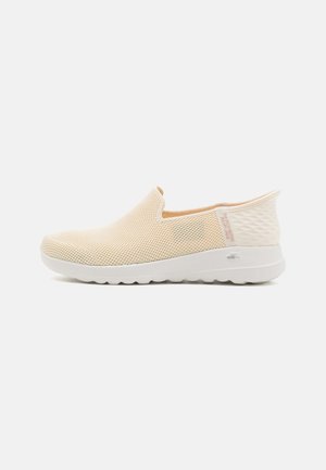 Zapatos slip-on color beige con parte superior de malla, suela de goma, puntera redonda y parte trasera texturizada. Presenta un branding mínimo en el talón. Diseño ligero.