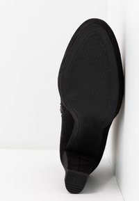 Semelle d'une botte noire à la cheville avec semelle en caoutchouc texturée et petit talon carré, positionnée debout contre un fond blanc d'angle.