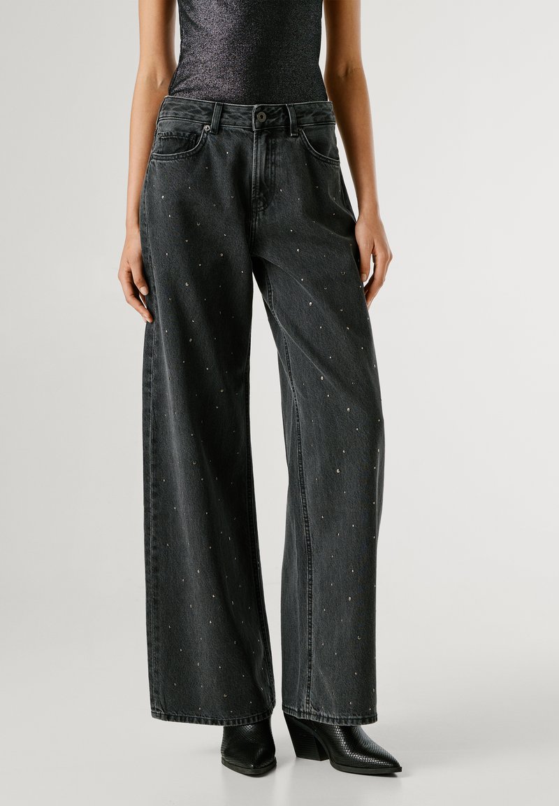 Plaša kāju, melna džinsu bikses ar metāla rotājumiem, vidēja augstuma jostasvieta, klasiskā piecu kabatu dizains un kontrastējoši šuvumi.