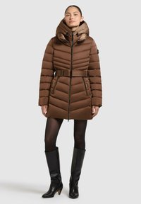 Braune Pufferjacke mit tailliertem Gürtel, Kapuze und Chevron-Steppung. Verfügt über Reißverschlusstaschen und wird mit knielangen schwarzen Stiefeln kombiniert.