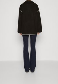 Czarna oversize kurtka z kołnierzem, z kontrastowym przeszyciem wzdłuż szwów, noszona na tle ciemnych dżinsów w fasonie dzwonów.