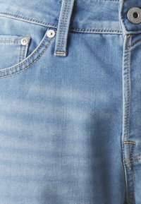 Jean en denim bleu clair avec une texture légèrement délavée, dotés de quincaillerie en laiton, d'une fermeture à bouton et de surpiqûres contrastées le long des coutures.