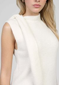Femme portant un haut en tricot blanc sans manches avec une écharpe blanche assortie drapée sur son épaule et des boucles d'oreilles créoles dorées.