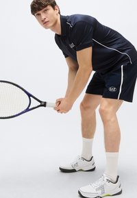 Temno modra polo majica z belimi poudarki, temne kratke hlače in bele superge s črnimi podplati. Drži teniški lopar, prikazuje športno atletsko oblačilo.