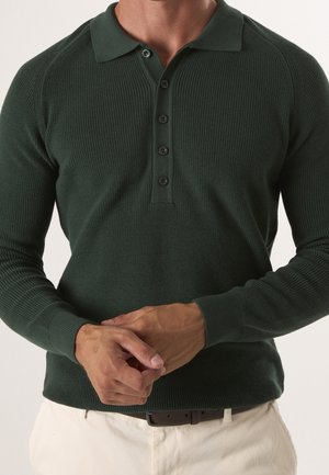 Homme portant un polo en maille vert foncé à manches longues avec quatre boutons, associé à un pantalon de couleur claire et une ceinture foncée, mains jointes.