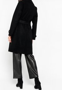 Czarny wełniany płaszcz z paskiem w talii, dużym kołnierzem i zapinanymi mankietami, do tego błyszczące czarne spodnie ze sztucznej skóry oraz czarne botki na obcasie.