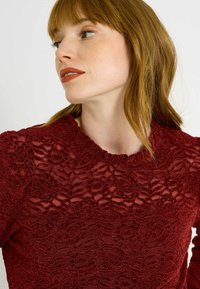 Haut en dentelle rouge avec motifs floraux, surface texturée, col montant et manches longues. Design détaillé et complexe sur tout le tissu.