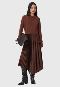 Brun beskuren tröja, brun plisserad midkjol, svarta knähöga stövlar, håller en mörk väska. Slät yta, lagerdesign och rika färger.