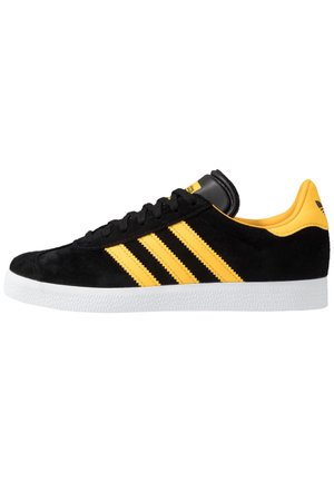 Czarny zamszowy sneaker z trzema żółtymi paskami, żółtymi akcentami na pięcie i języku oraz białą teksturowaną podeszwą.