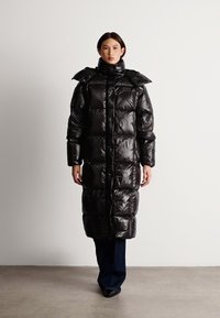 Geacă puffer neagră cu finisaj lucios, siluetă lungă, guler supradimensionat și textură matlasată. Are un fermoar frontal și detaliu cu logo.