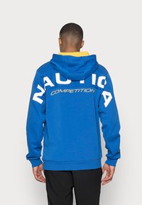 Blå huvtröja med vit "NAUTICA" text på ryggen och "COMPETITION" i gult. Ribbad ärm- och nederkant. Mjuk textur.