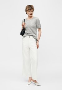 Kobieta ubrana w szary, krótki sweter z krótkim rękawem, białe spodnie o szerokich nogawkach, białe, spiczaste szpilki i trzymająca małą czarną torebkę na ramię.