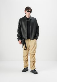 Jachetă bomber din piele neagră, tricou negru, pantaloni khaki cu detalii plisate, ochelari de soare și adidași negri. Detalii notabile includ curele pe pantaloni.