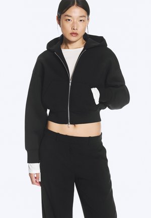 Jeune femme portant un sweat à capuche court noir zippé, un chemisier blanc à manches longues en dessous et un pantalon noir, debout avec une main dans la poche.