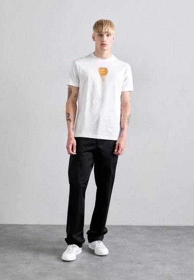 Man with short blonde hair wearing a white Versace t-shirt, black pants, white sneakers, standing against a light gray wall.