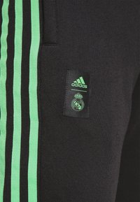 Sort stof med tre vertikale neongrønne striber og et mærke, der viser grønne Adidas- og Real Madrid-logoer.