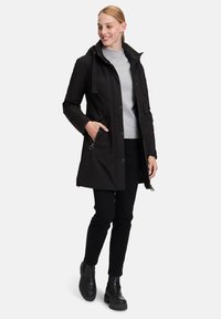 Schwarze wasserdichte Jacke mit hohem Kragen, sichtbaren Knöpfen, zwei Seitentaschen und Kapuze mit Kordelzug. Getragen mit schwarzen Hosen und Stiefeln.