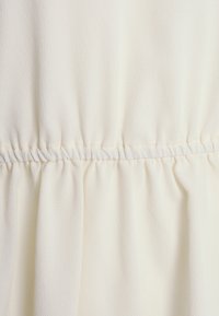 Tessuto color crema con una superficie leggermente strutturata, arricciato in vita con un elastico per aumentare il drappeggio e la pienezza.