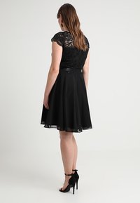 Donna in un abito nero al ginocchio con top in pizzo e gonna trasparente, che indossa scarpe col tacco alto nere con cinturino alla caviglia, di spalle.