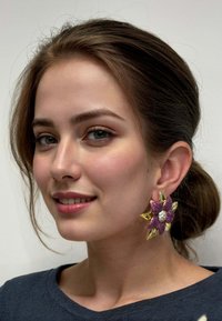 Gold floral earrings featuring pink gemstones and a central clear stone, set against a textured surface, worn by a model with a neutral top.