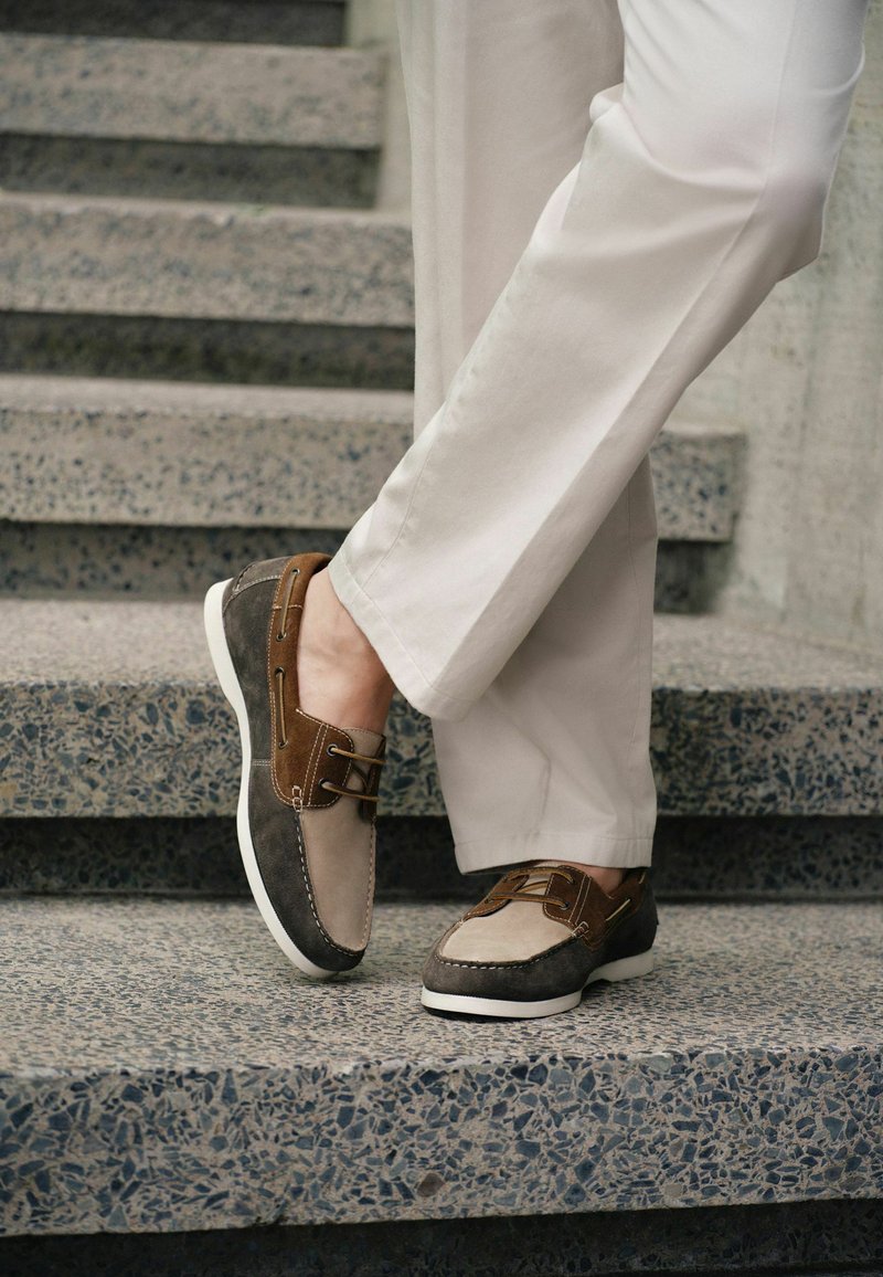 Personne portant des chaussures bateau en cuir suédé beige et marron et un pantalon beige clair, debout sur des escaliers en pierre tachetée.