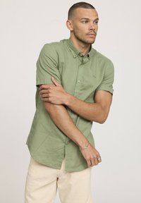 Chemise à manches courtes vert clair avec boutons, présentant une texture subtile, dotée d'un col évasé, d'une poche avant et d'un ourlet arrondi.