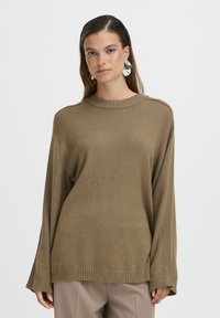Suéter de punto marrón con un cuello alto, corte holgado y puños y dobladillo acanalados. El modelo lleva grandes pendientes de plata. Diseño minimalista, color neutro.