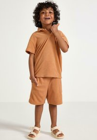 Terra-cotta-farvet, kortærmet hoodie og shorts sæt i en afslappet pasform, med en tekstureret overflade og sidelommer. Båret med sandaler.