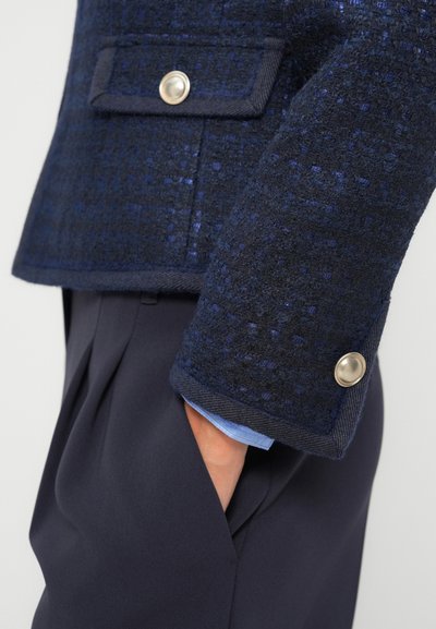 Giacca blu marinato in tessuto strutturato con una combinazione di materiali, caratterizzata da un colletto a punta e un dettaglio con bottone argentato sulla manica. Abbinata a pantaloni scuri.