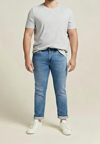 Homme portant une chemise à manches courtes gris clair, un jean bleu retroussé et des baskets blanches, debout devant un fond beige uni.