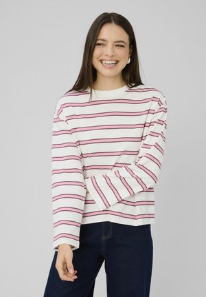 Jeune femme souriante, portant une chemise blanche à manches longues avec des rayures horizontales roses et un jean bleu foncé, debout devant un fond uni.