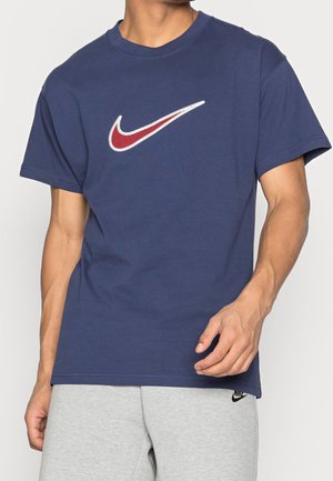 Mężczyzna w granatowej koszulce z czerwono-białym logo Nike oraz jasnoszarych spodniach dresowych z małym czarnym logo Nike.