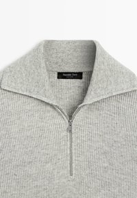 Hellgrauer Pullover mit einem gerippten Muster, ausgestattet mit einem Reißverschlusskragen und einem Etikett innen. Hergestellt aus weichem Material für hohen Komfort.