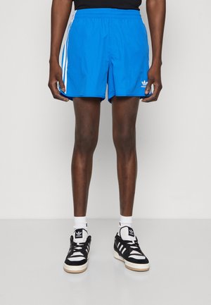 Man draagt felblauwe Adidas-shorts met witte strepen, zwart-witte Adidas-sneakers en witte sokken tegen een effen achtergrond.