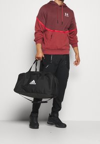 Schwarze Sporttasche mit weißem Adidas-Logo, getragen mit einer kastanienroten Kapuzenjacke mit rotem Streifen, kombiniert mit schwarzen Sporthosen und Schuhen.