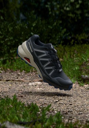 Scarpa da trail running Salomon nera e grigia in aria sopra un sentiero ghiaioso con vegetazione verde sullo sfondo.