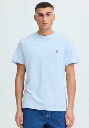 Młody mężczyzna ubrany w jasnoniebieską koszulkę z okrągłym dekoltem z małym logo pszczoły na lewej piersi oraz niebieskie jeansy, stojący na jednolitym tle.