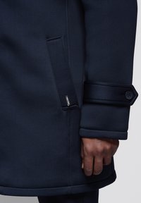 Navy blue coat with a smooth texture, featuring a side pocket and buttoned cuff detail. The fabric is structured and tailored for a fitted look.