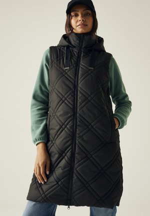 Femme portant une longue veste matelassée noire sans manches avec une capuche, par-dessus une polaire verte et un jean bleu.