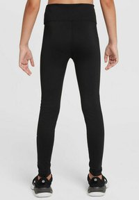 Leggings noirs avec une taille haute, texture lisse et une coupe ajustée. Portés avec des chaussures de sport noires à semelle blanche.