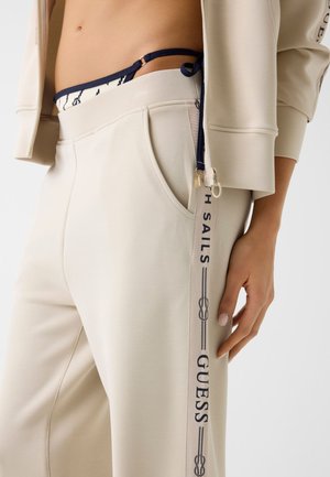 Pantaloni della tuta beige con tasca frontale e strisce laterali con la scritta "H Sails Guess", indossati con una giacca beige corta e un costume bikini blu navy a fantasia.