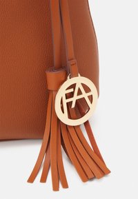 Brun tekstureret lædertaskehjørne med matchende læderkvaster og et rundt guldfarvet metalvedhæng med udskårne bogstaver FA.