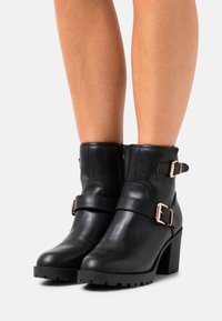 Botas negras hasta el tobillo de cuero con un tacón robusto, que cuentan con doble hebilla lateral y cierre de cremallera, suela texturizada para un mejor grip.