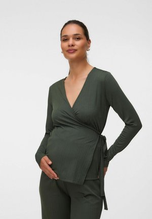 Gravid kvinde iført en mørkegrøn omslagstop med lange ærmer og matchende bukser, blidt holdende om sin mave, smilende blidt.