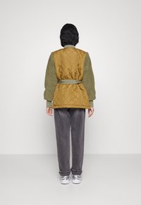 Quiltad senapsgul och grön jacka med bältesmidja, med en texturerad yta. Buren med lösa grå byxor och vita sneakers.