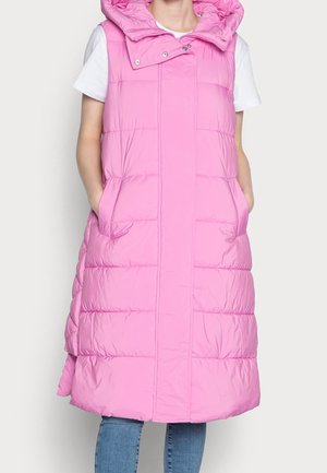 Femme portant un long gilet matelassé rose sans manches avec des poches avant, par-dessus un t-shirt blanc à manches courtes et un jean bleu, les mains dans les poches.