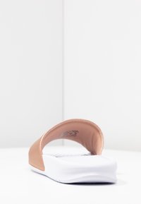 Sandale à enfiler avec une semelle blanche coussinée et une large bride beige sur le pied, vue d’un angle arrière bas sur une surface blanche.